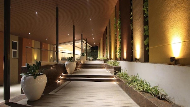 A corridor with wooden flooring, large potted plants, and illuminated wall sconces. - Icon Business by Bhagini  (Bengaluru)