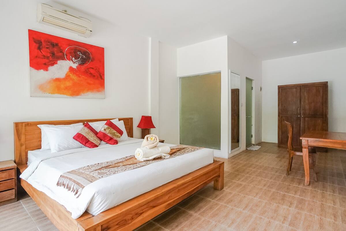 A neatly made bed with white and red pillows, a wooden headboard, and a red abstract painting above it.