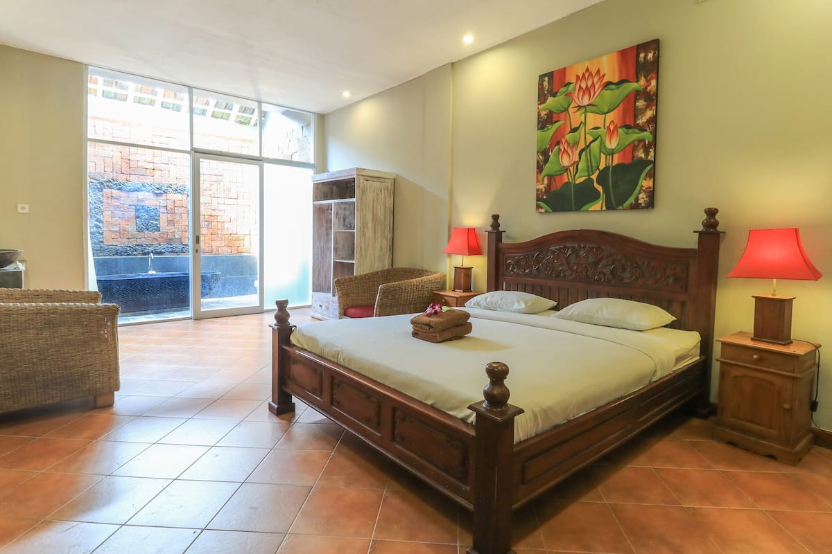 A spacious bedroom with a large bed, a wooden wardrobe, a wicker chair, and a view of a brick wall through a sliding glass door.