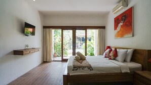 A bedroom with a bed, a TV, a painting, and a sliding glass door leading to a garden. - D'uma Residence (Denpasar)
