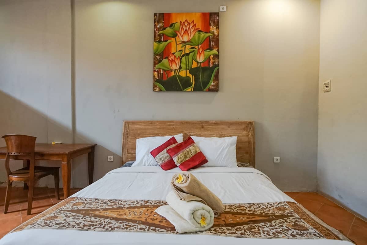 A hotel room with a wooden bed, white bedding, a patterned rug, a wooden chair, and a painting of lotus flowers on the wall.