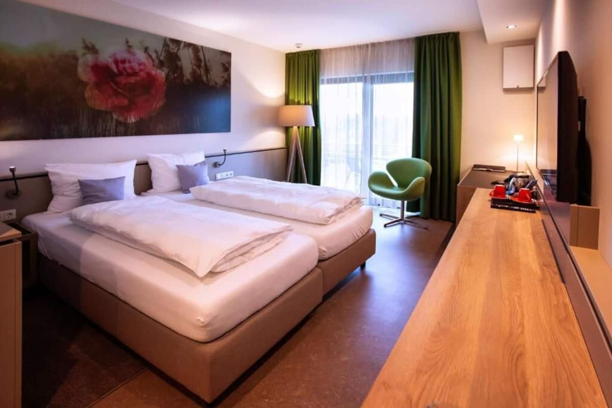 A hotel room with a double bed, a desk, a chair, a large painting, and a window with curtains.
