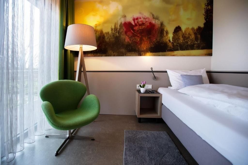 A hotel room with a bed, a green chair, a nightstand, a lamp, and a painting on the wall.