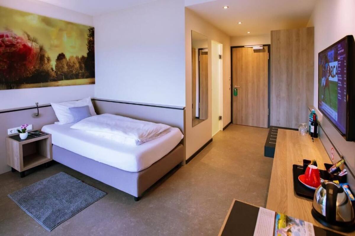 A modern hotel room with a large bed, a desk with a coffee maker, a TV, and a painting on the wall.