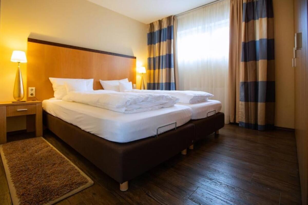 A hotel room with a large bed, two bedside tables, a lamp, and a view of the outdoors through curtains.