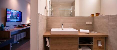 A modern bathroom with a wooden vanity, a large mirror, and a television mounted on the wall.