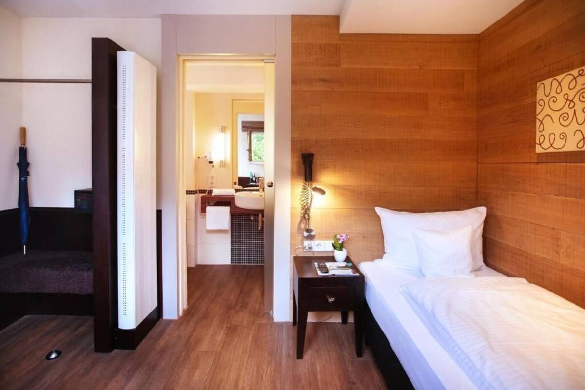 A hotel room with a wooden headboard, a bed with white linens, a nightstand with a lamp, and a view of the bathroom and hallway.
