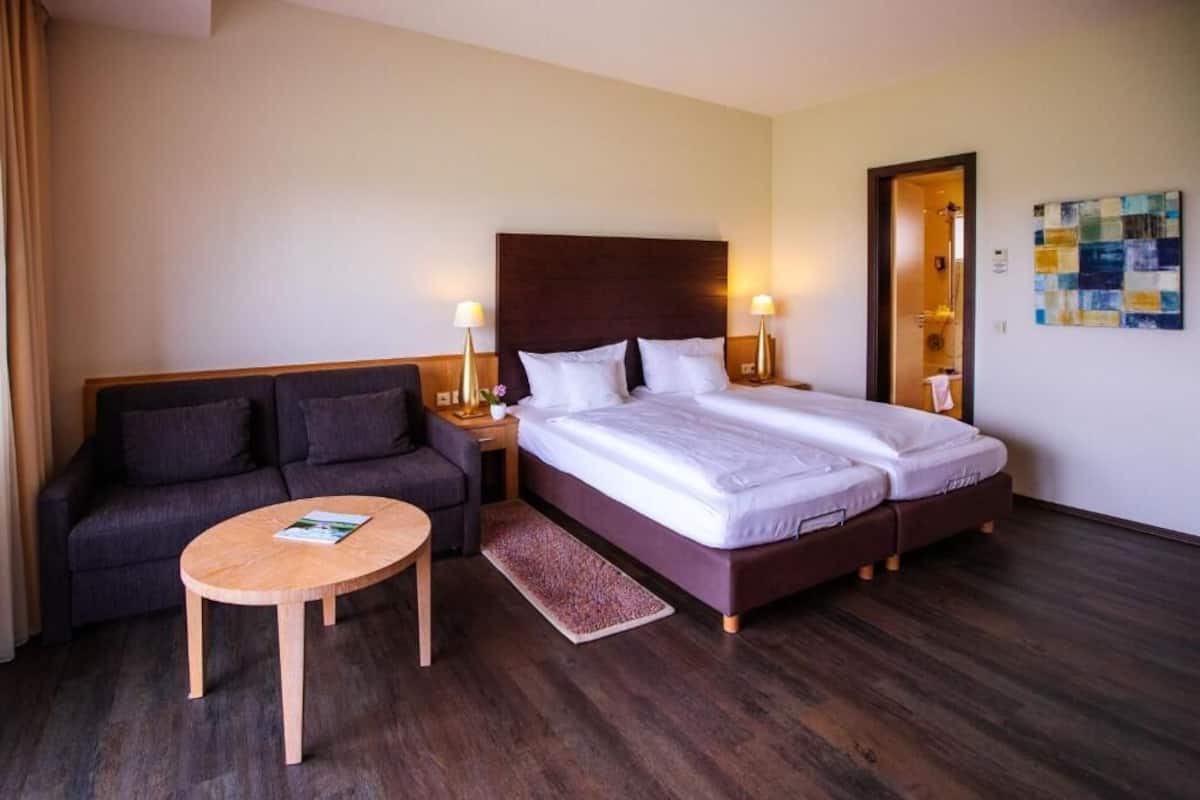 A hotel room with a double bed, a sofa, a small wooden table, and two bedside lamps.