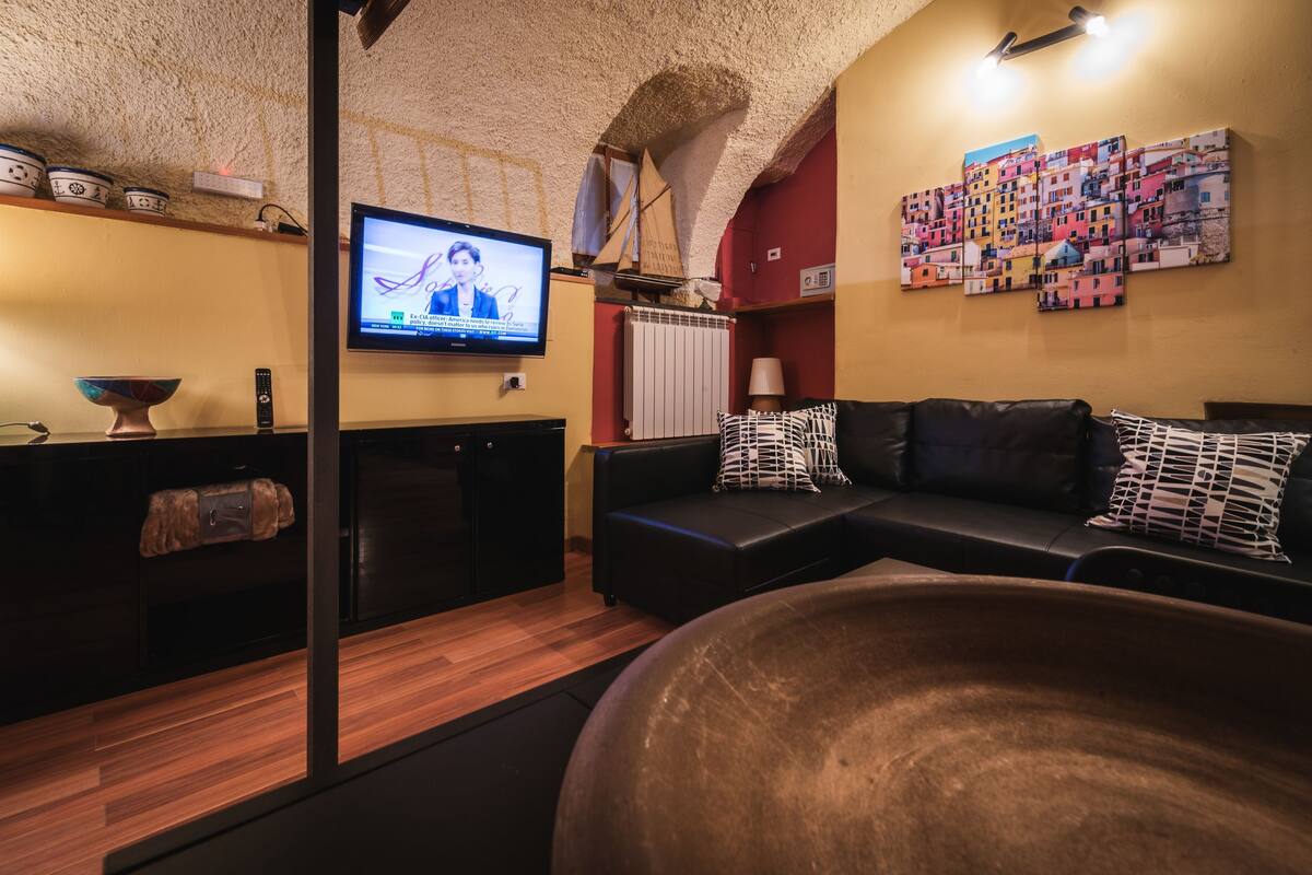 A modern living room with a black leather sofa, a large wooden bowl, a television, and a colorful wall art piece.