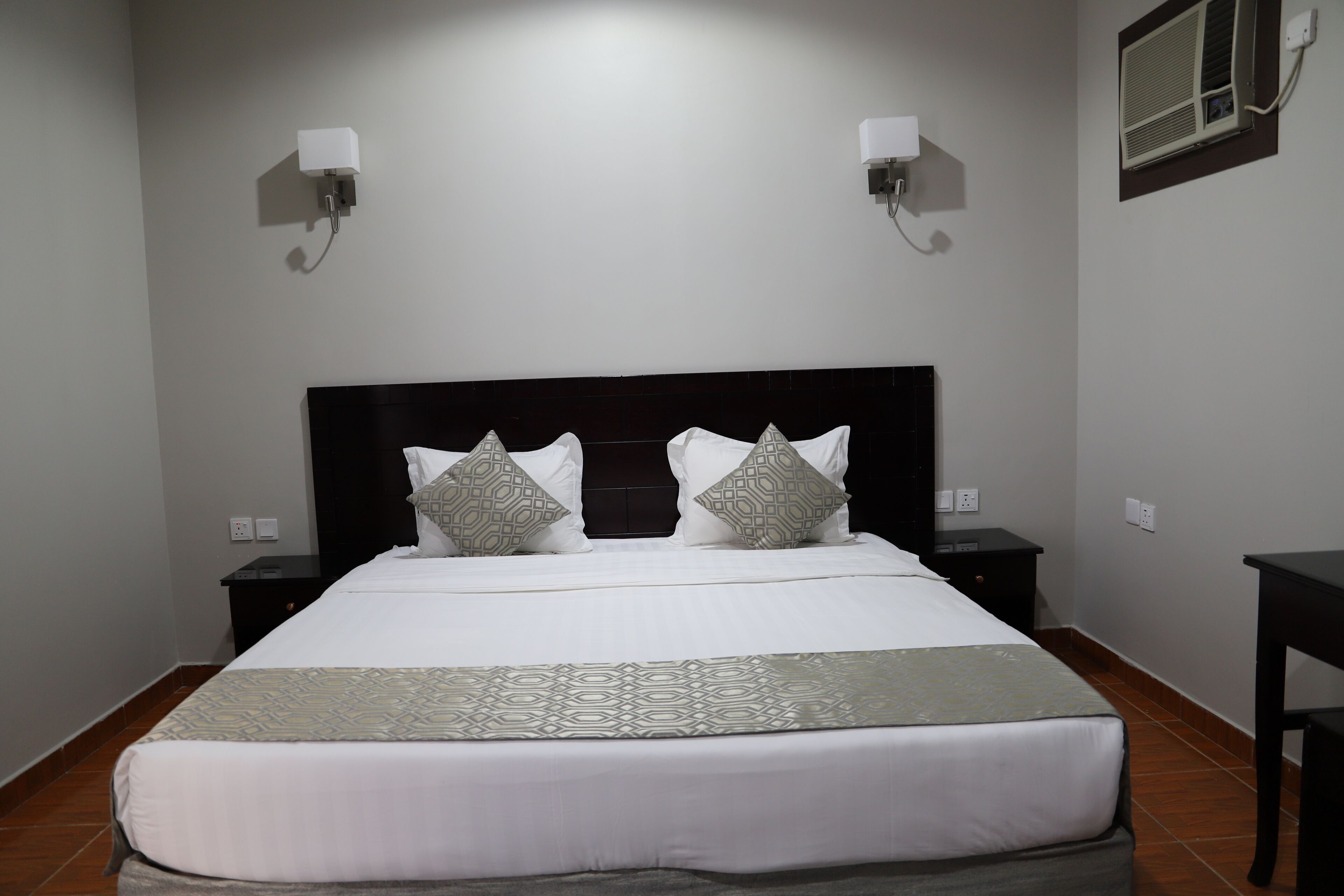 A neatly made bed with a patterned bedspread, two bedside tables with lamps, and a wall-mounted air conditioning unit.