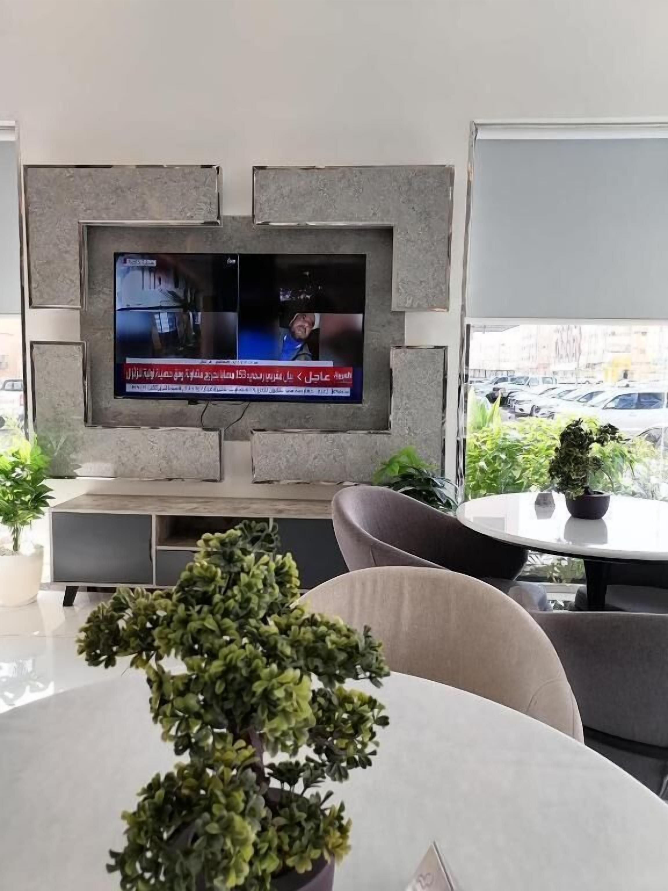 A modern living room with a flat-screen TV mounted on a stone wall, a round dining table with chairs, and potted plants.