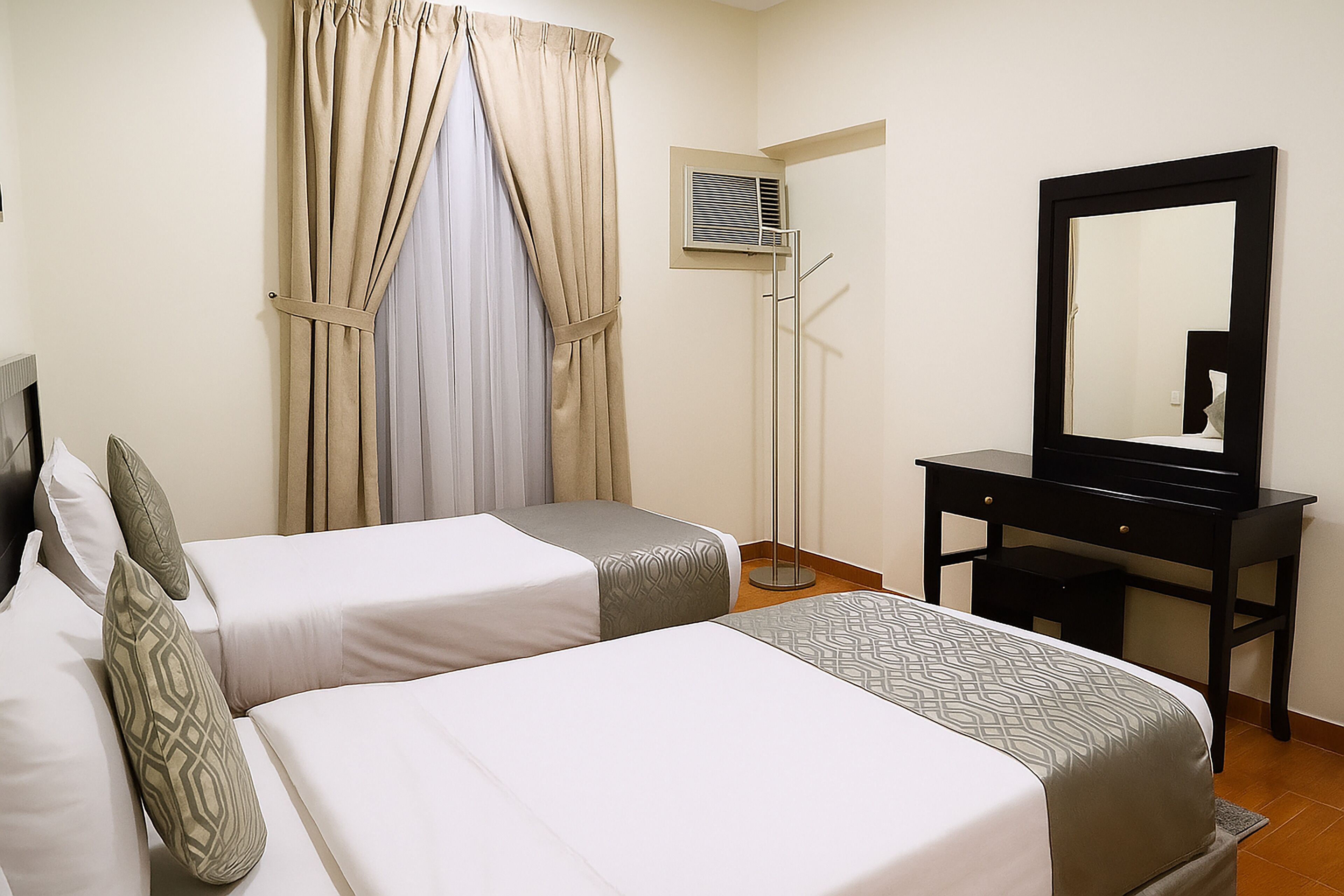 A hotel room with two beds, a desk, a mirror, and an air conditioning unit.