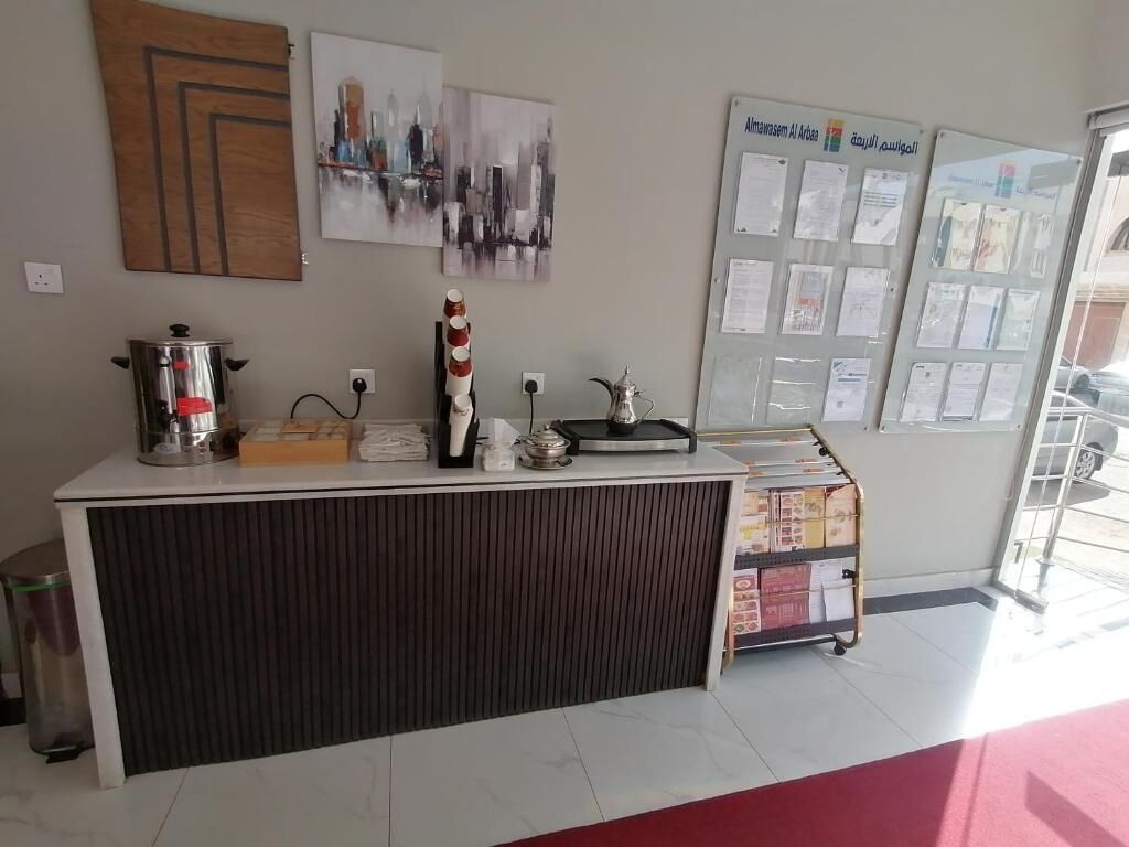 Una recepción con una máquina de café, un hervidor y un mueble bajo. Hay cuadros en la pared y un tablón de anuncios con papeles.