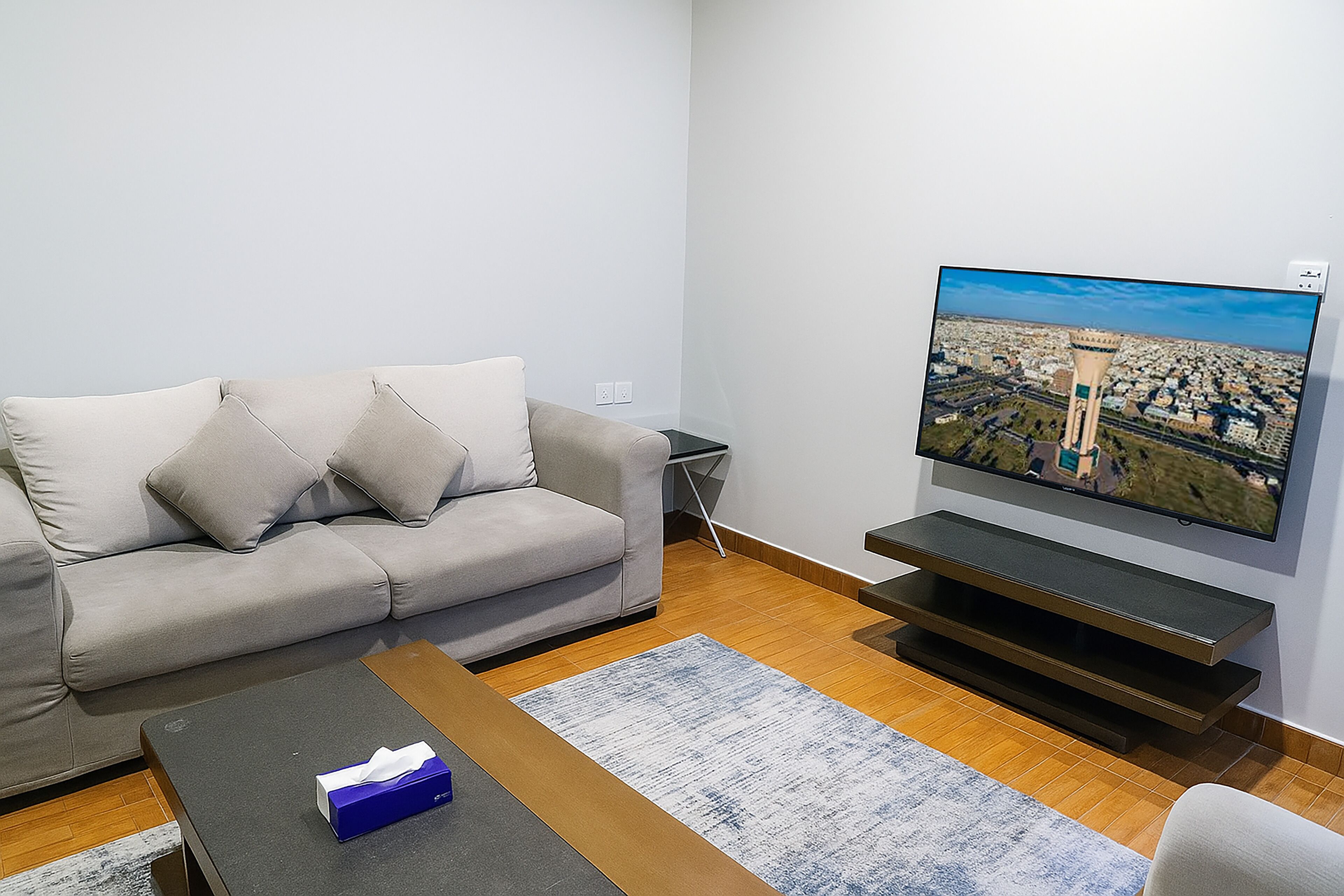 A living room with a grey sofa, a coffee table with a tissue box, a flat-screen TV on a stand, and a wooden floor.
