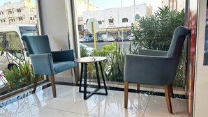 A modern indoor seating area with two blue chairs and a small table, large windows with a view of the street and buildings, and a tiled floor. - Al Mawasim Al Arbaeah Serviced Apartments (Tabuk)