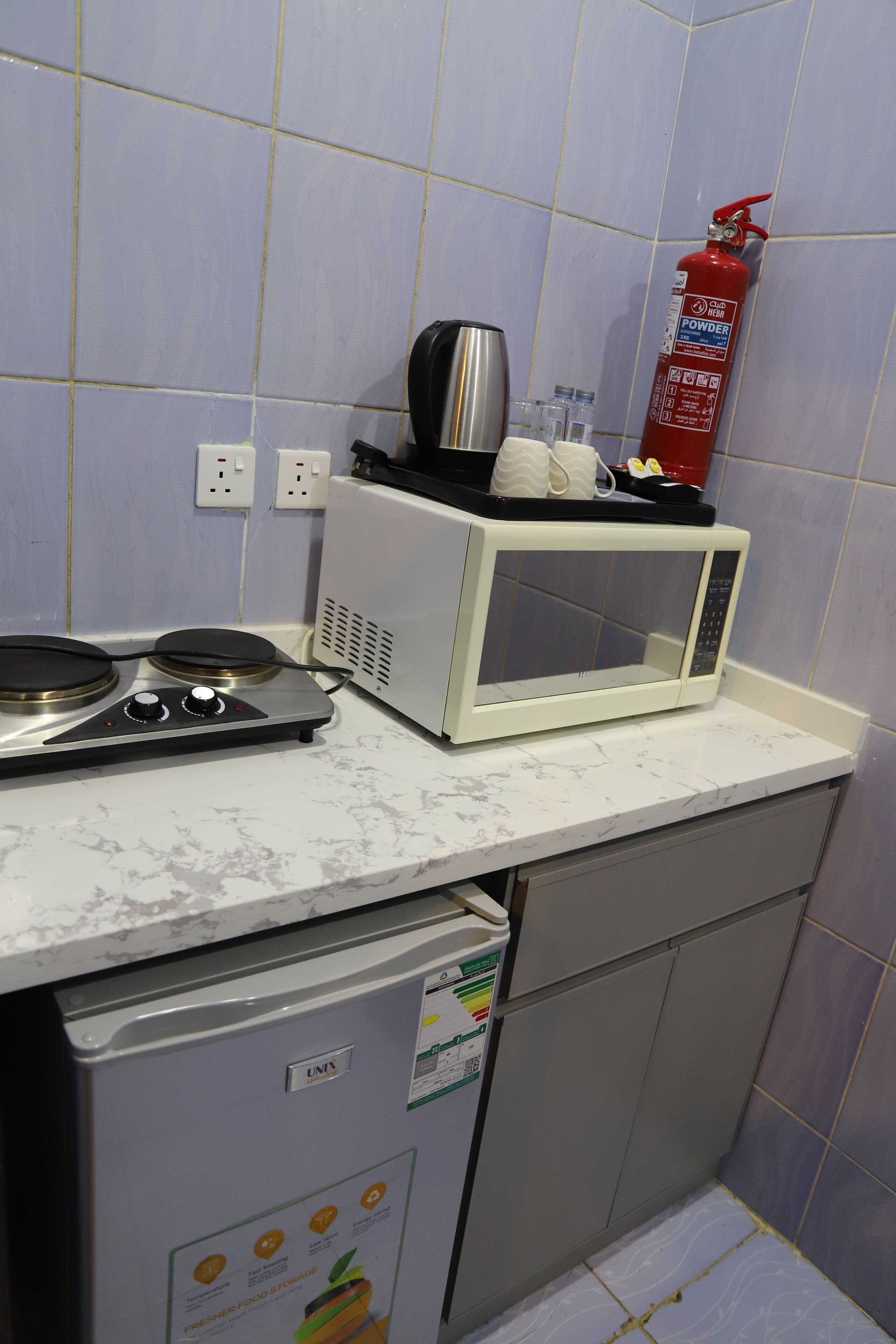 Una cocina equipada con microondas, cafetera, extintor y un refrigerador pequeño.