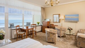 Habitación de hotel con un ventanal amplio que ofrece vistas al mar, una cama, un escritorio con una silla, dos sillones, una mesita con un florero y un candelabro.
