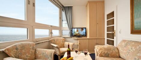Una cabaña acogedora con un ventanal grande que ofrece vistas al mar, un televisor y una mesa con fruta y copas de vino.