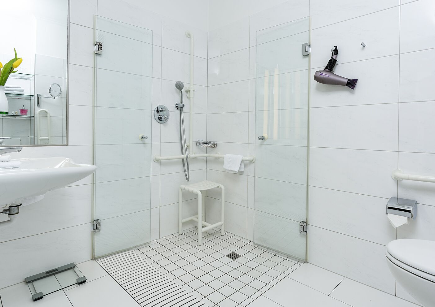 Un baño moderno con ducha envuelta en vidrio, azulejos blancos, un lavamanos, inodoro y un secador de pelo montado en la pared.