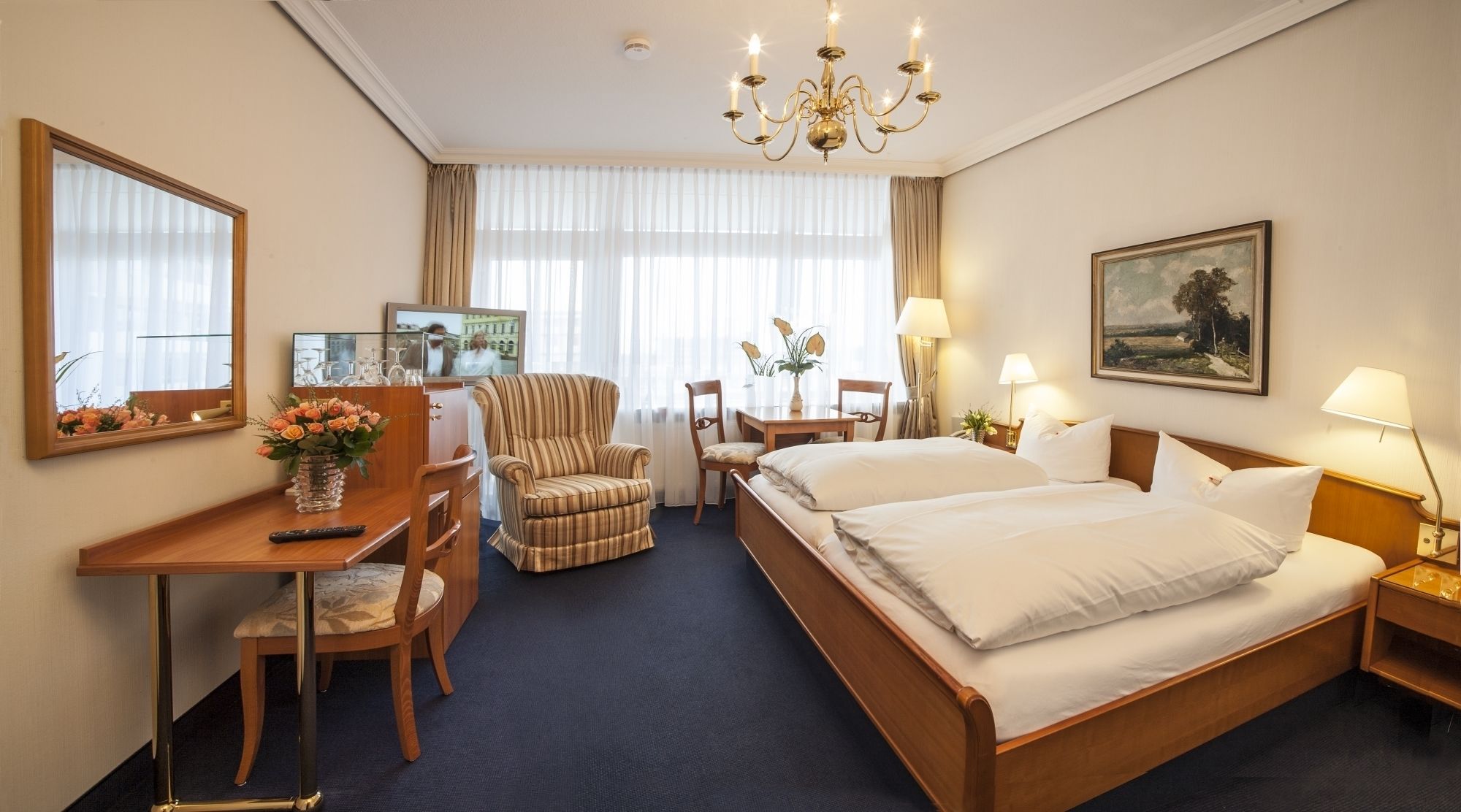 A hotel room with a bed, desk, chair, TV, and a chandelier.