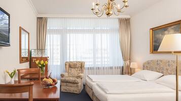 Habitación de hotel con cama, escritorio con silla, sillón pequeño, candelabro y un cuadro en la pared.