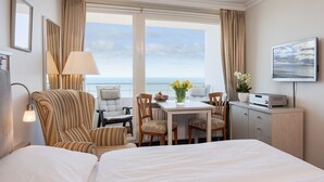 Habitación de hotel con una cama grande, una mesa de comedor con sillas, un televisor y vistas al mar.