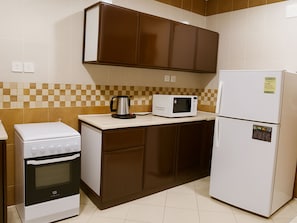 A compact kitchen with a stove, microwave, and refrigerator, featuring brown cabinets and checkered tile backsplash. - Binayat Saraya Serviced Apartments (Tabuk)