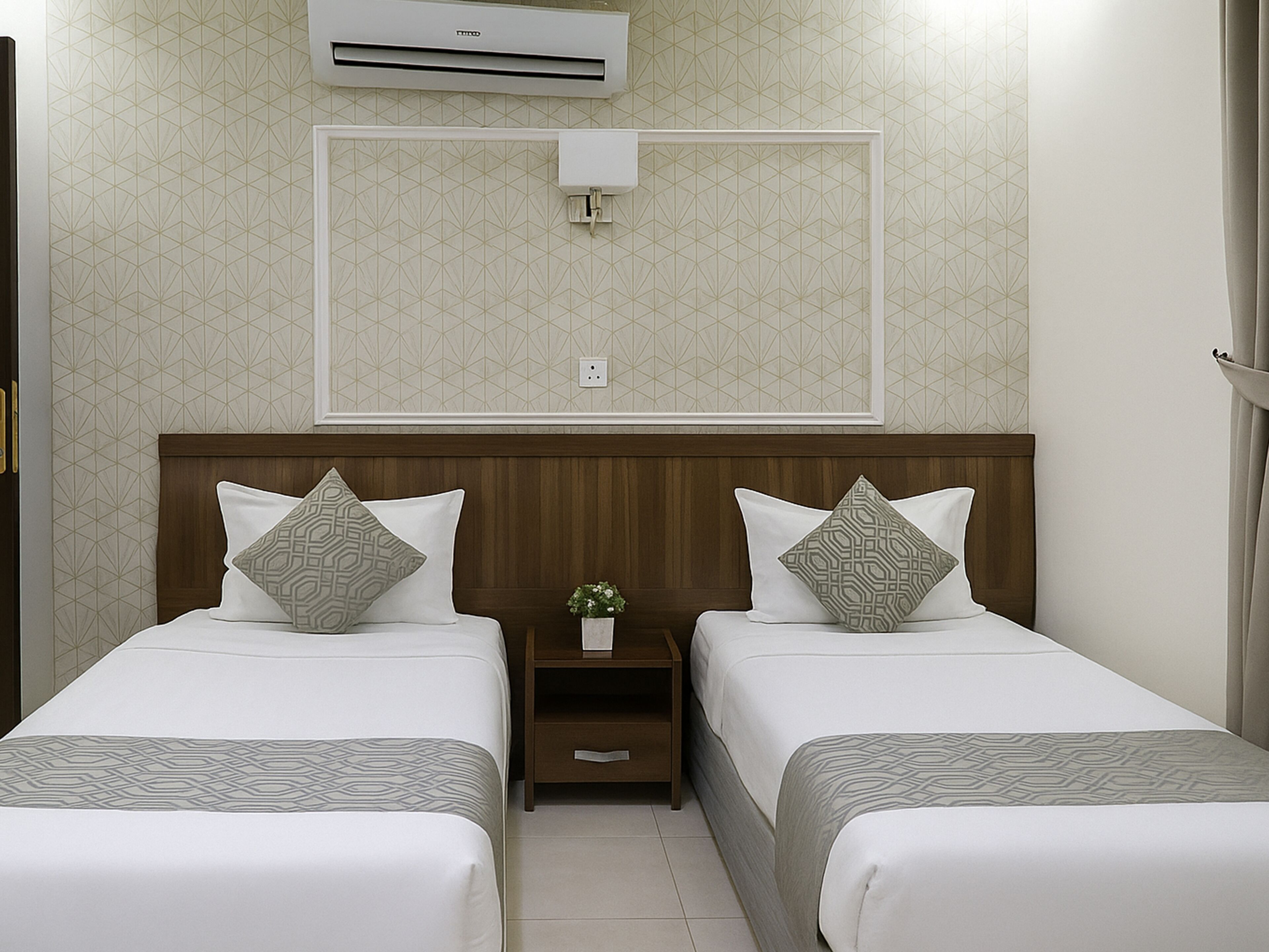A hotel room with two beds, a nightstand, a small potted plant, and an air conditioning unit on the wall.