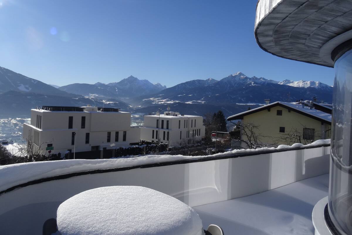 A snow-covered balcony with a view of snow-capped mountains and buildings in the background.