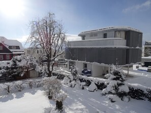 En modern byggnad med ett platt tak och stora fönster, omgiven av snötäckta träd och buskar.