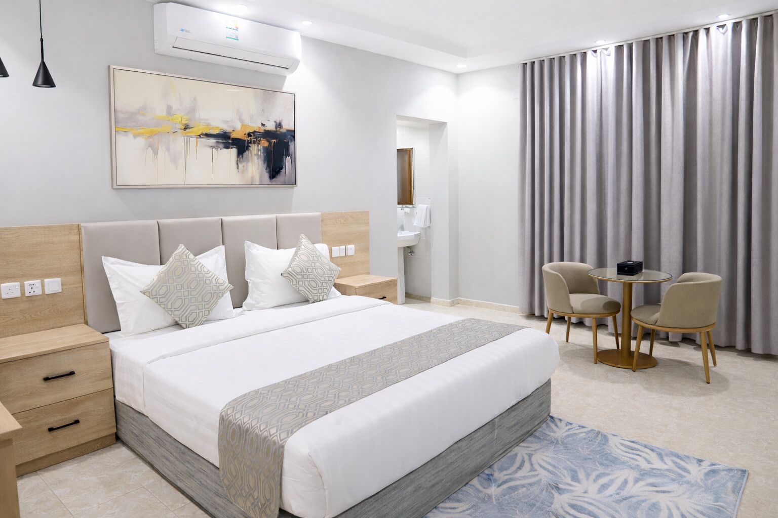 Una habitación de hotel moderna con una cama grande, mesitas de noche, un escritorio con sillas, un baño y un ventanal con cortinas.