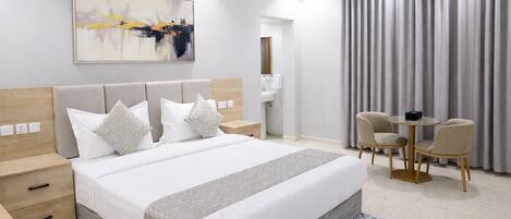 Una habitación de hotel moderna con una cama grande, mesitas de noche, un escritorio con sillas, un baño y un ventanal con cortinas.