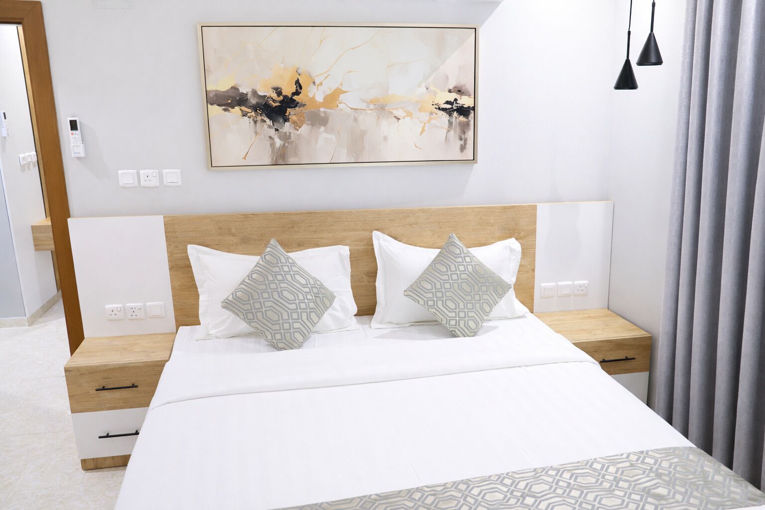 A hotel room with a large bed, two bedside tables, a nightstand, a wall-mounted artwork, and a grey curtain.