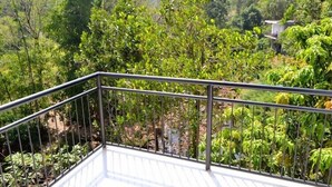 Een balkon met een metalen hekwerk, met uitzicht op een weelderig groen landschap met dichte begroeiing en een gebouw in de verte.