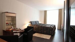 A hotel room with a bed, a desk with a chair, a small table with items, a lamp, and a TV.