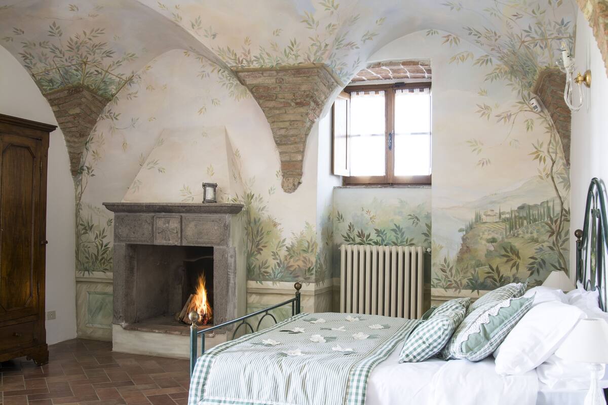 A bedroom with a fireplace, a bed with checkered bedding, and a mural on the walls.