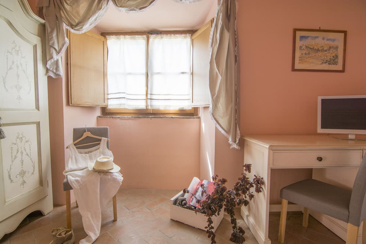 A bedroom with a bed, a desk, a chair, a window with curtains, a television, and a small plant.