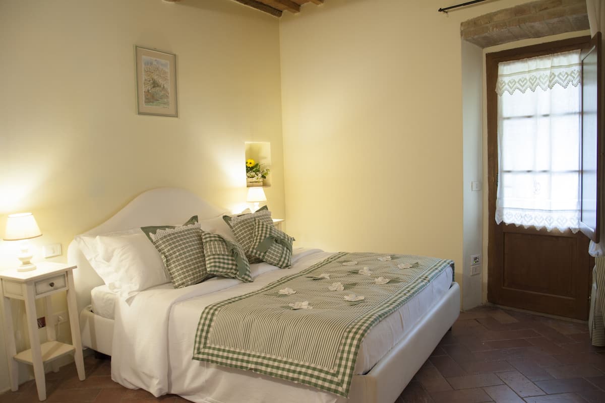 A neatly made bed with a checkered bedspread, a bedside table with a lamp, and a small wall-mounted shelf with flowers.