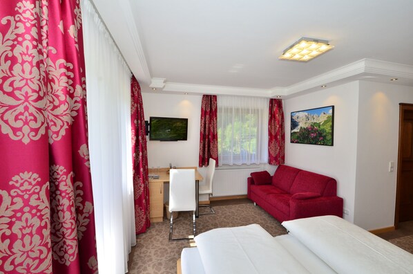 Quarto de hotel com cama, escrivaninha, cadeira, um sofá vermelho e duas pinturas na parede.