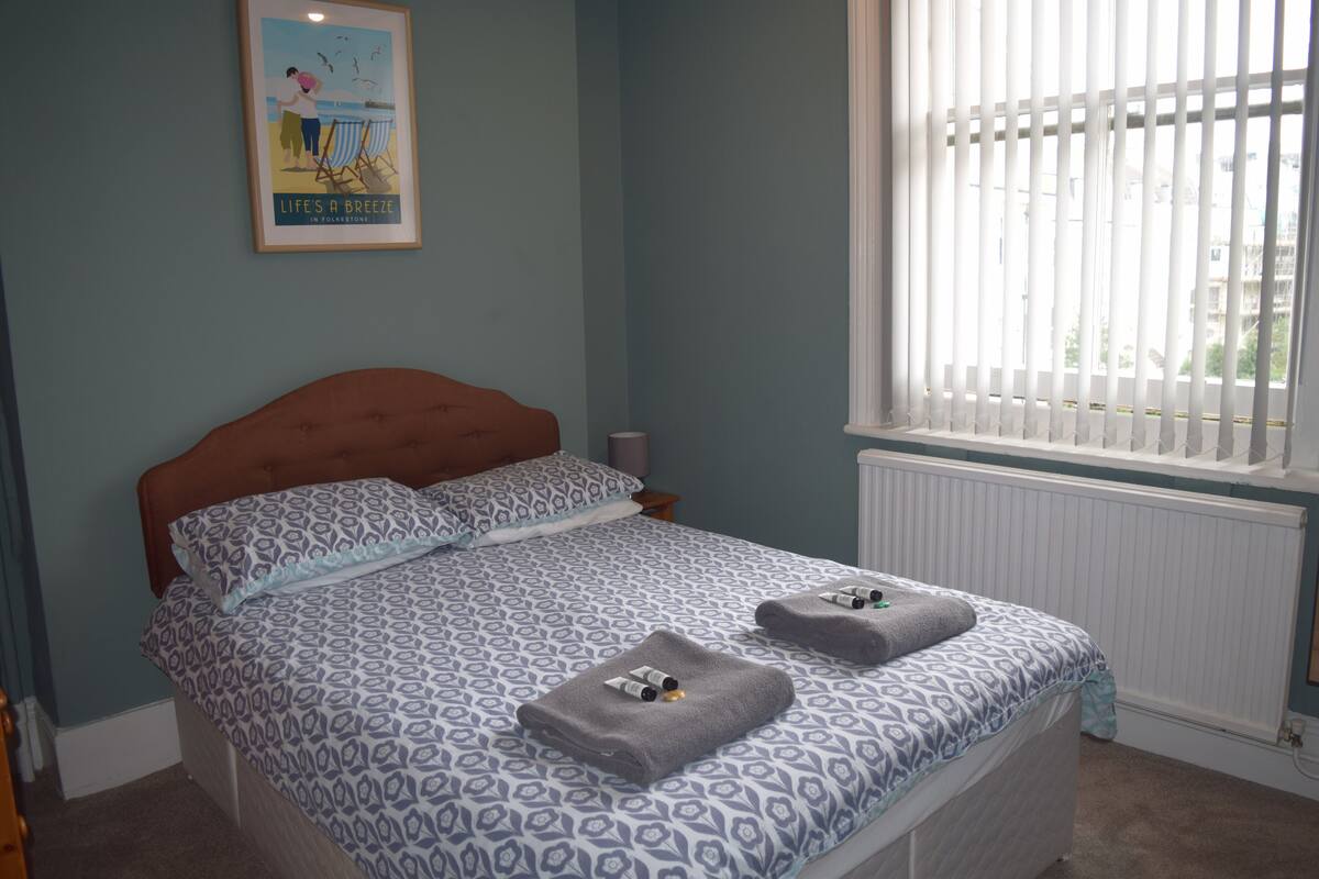 A bedroom with a patterned bedspread, two towels on the bed, a framed picture on the wall, and a window with blinds.