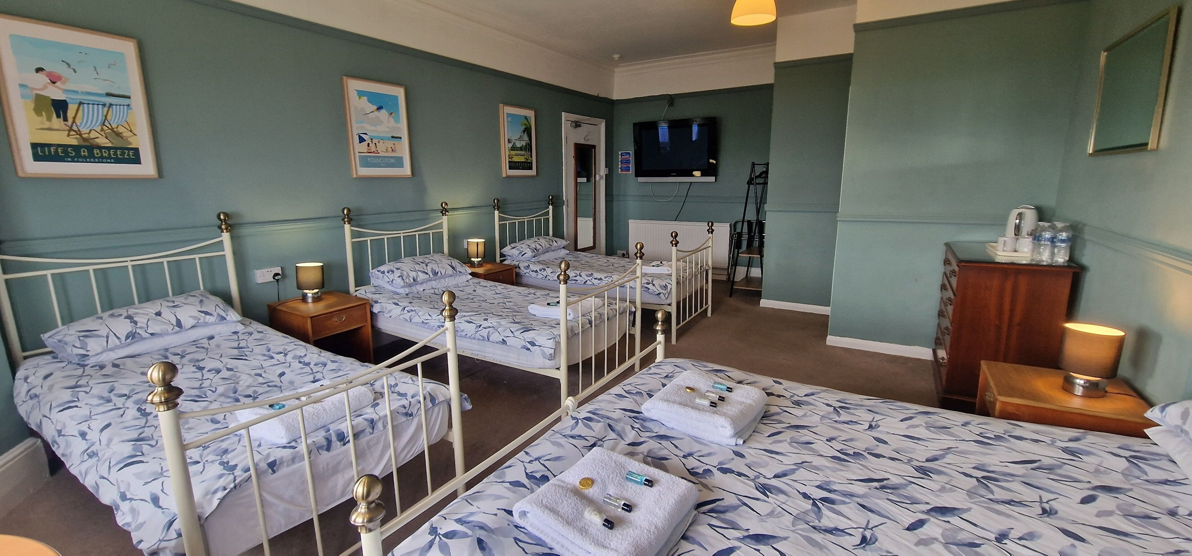 A hotel room with four single beds, a TV, a wooden dresser, and a small table with a lamp.