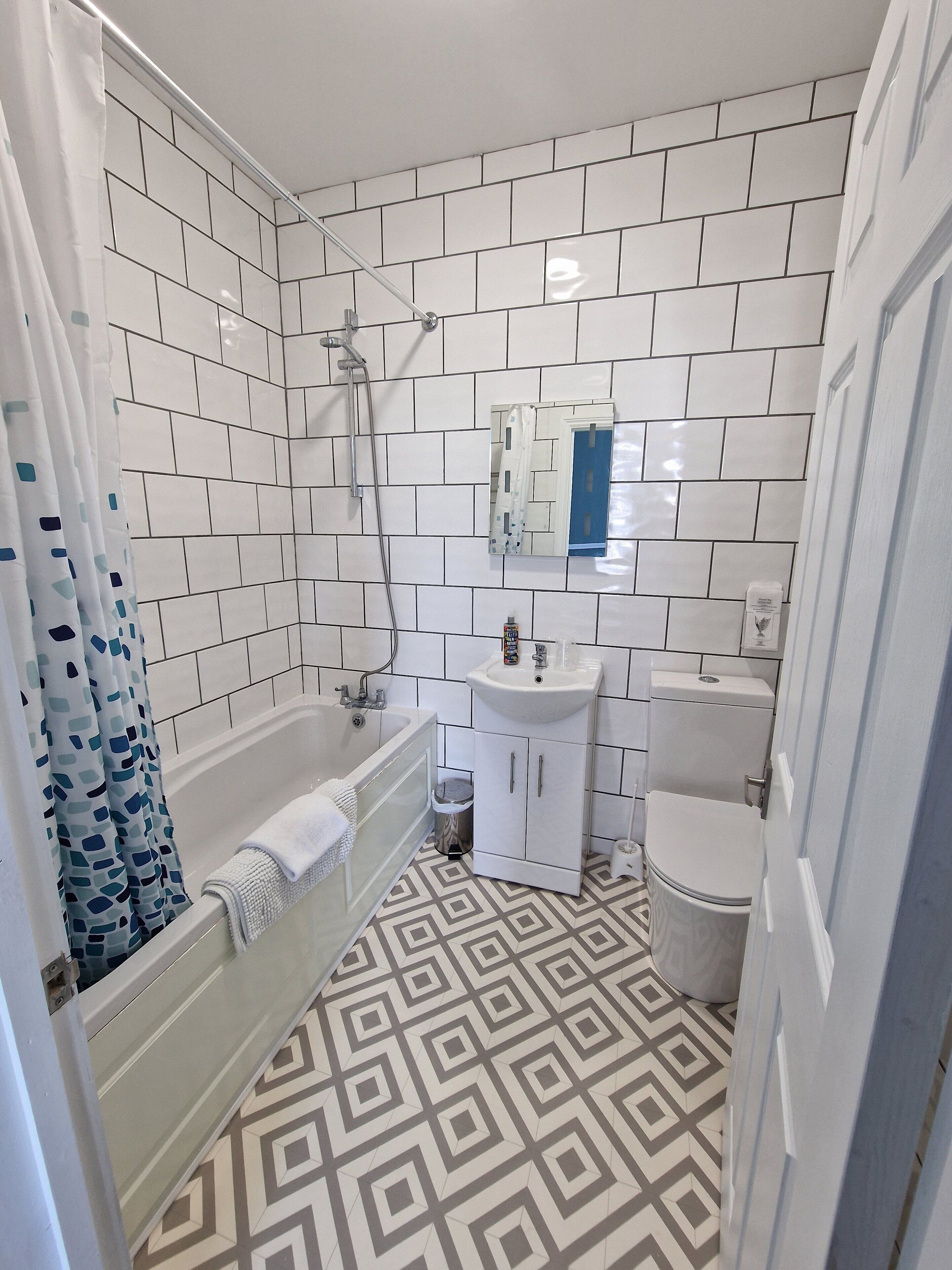 Un baño con paredes de azulejos blancos, piso estampado, bañera, lavamanos y retrete.