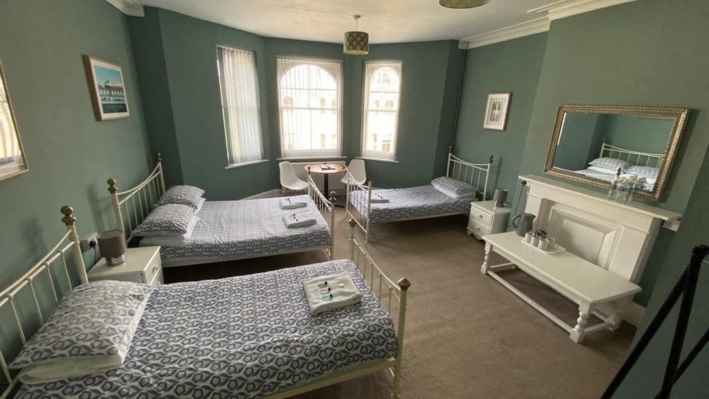 A room with three single beds, a small round table, a white bench, and a mirror on a dresser.