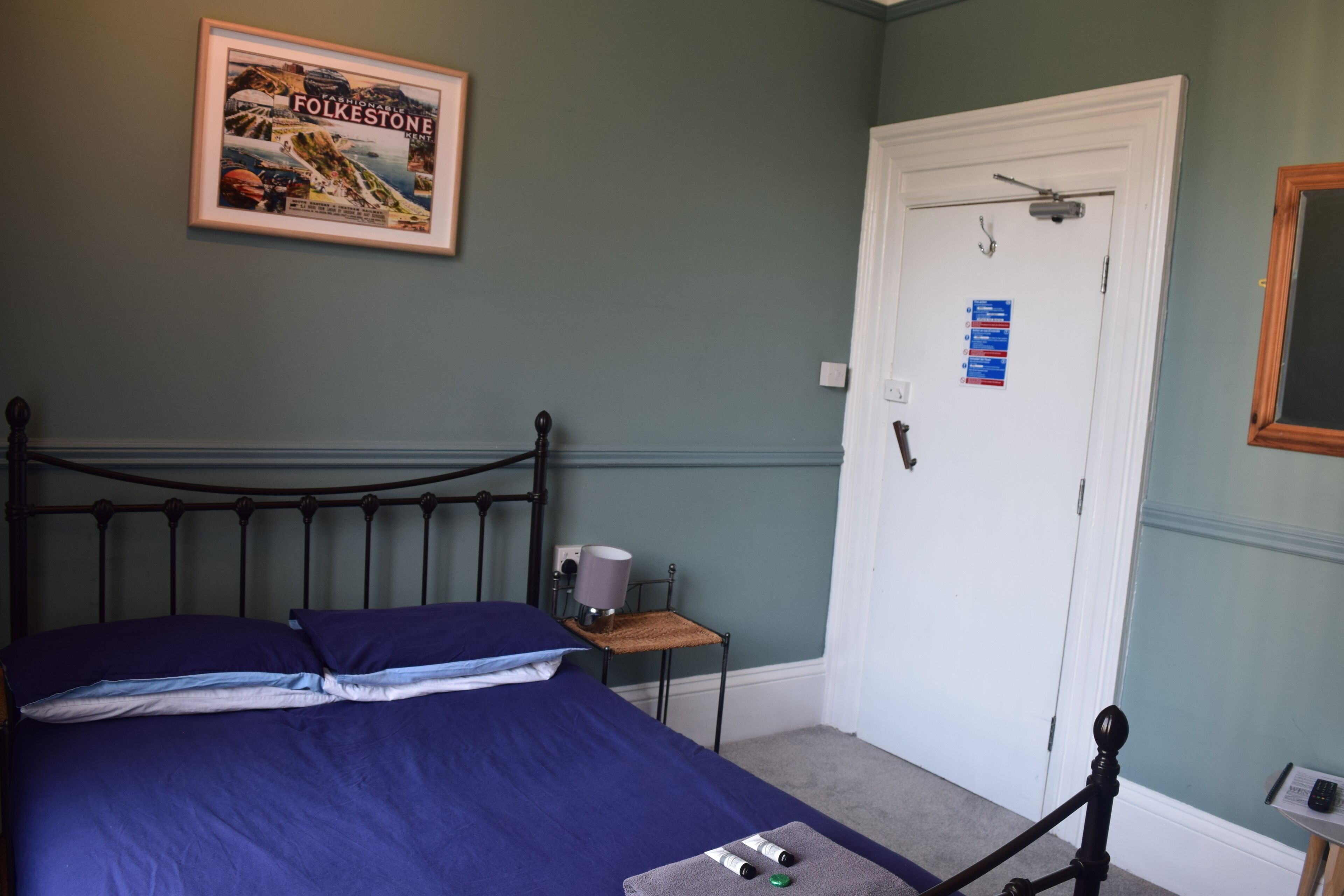 A bedroom with a bed, a framed picture on the wall, a door, and a small table with a lamp.