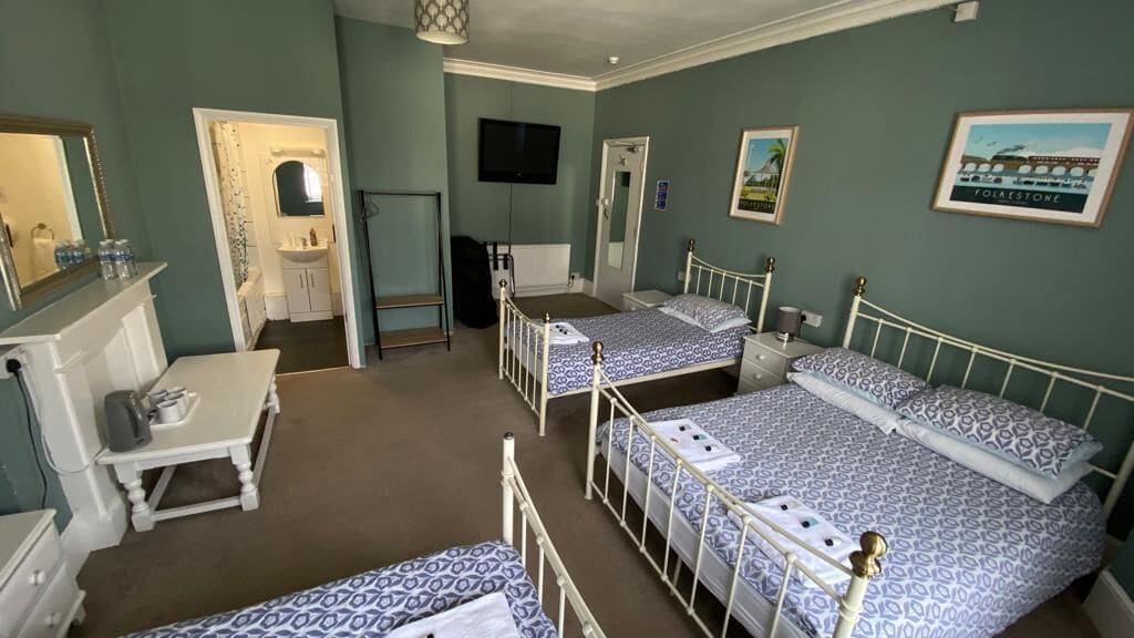 A room with three beds, a desk, a mirror, a television, and framed pictures on the wall.