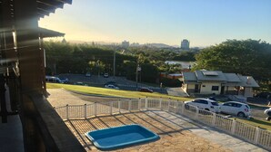 A swimming pool with a tiled deck, surrounded by a white fence, and a view of a road with cars and buildings in the background. - Hotel Panorâmico (Penha)