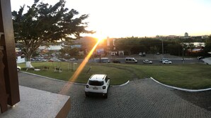 A parking area with a white car, a grassy area, and buildings in the background. - Hotel Panorâmico (Penha)