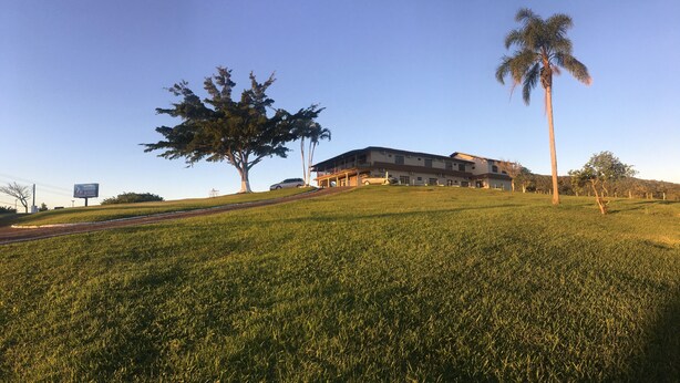 A hotel with a large tree and a palm tree in front, a grassy hill, and a clear sky. - Hotel Panorâmico (Penha)