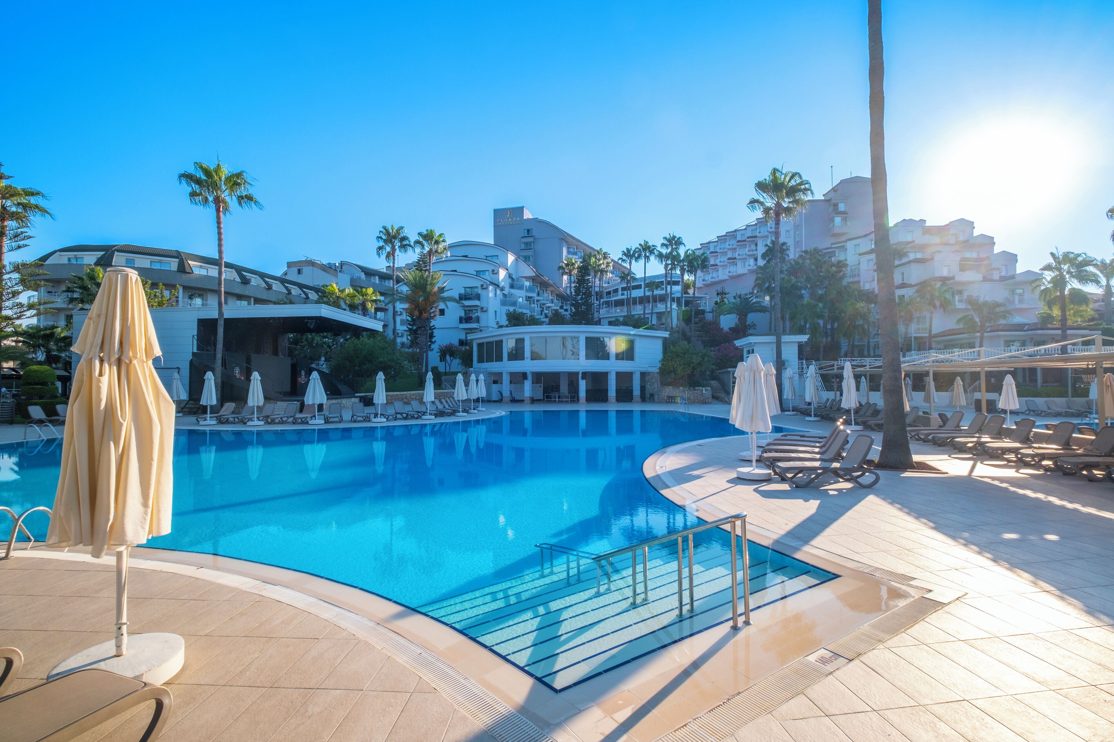 A swimming pool with lounge chairs and umbrellas, surrounded by palm trees and modern buildings.