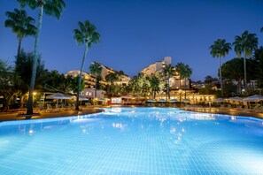 Una piscina con sillas de descanso y sombrillas, rodeada de palmeras y edificios de hotel al anochecer.